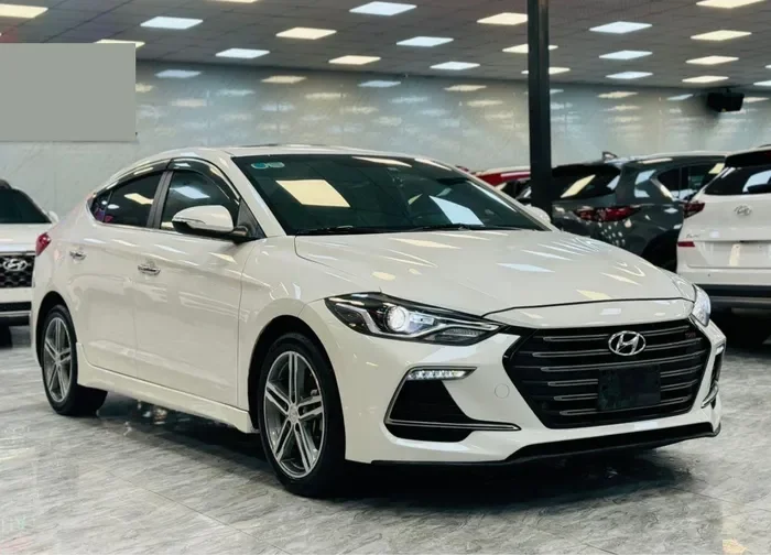 Hyundai Elantra cũ màu trắng đỗ trên đường phố, phía trước tòa nhà hiện đại.